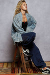 Person sitting on a wooden stool wearing a denim jacket and blue jeans against a gray wall.