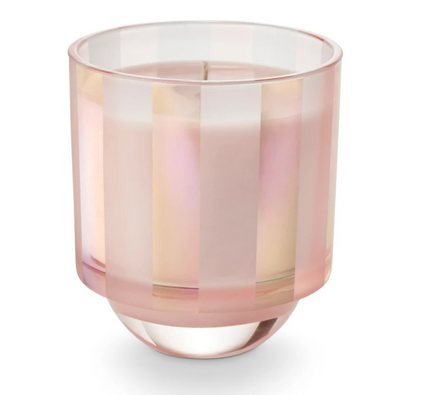 A multicolored striped glass candle, with a pinkish hue at the bottom, sitting inside a box with shiny, tinsel-like details.