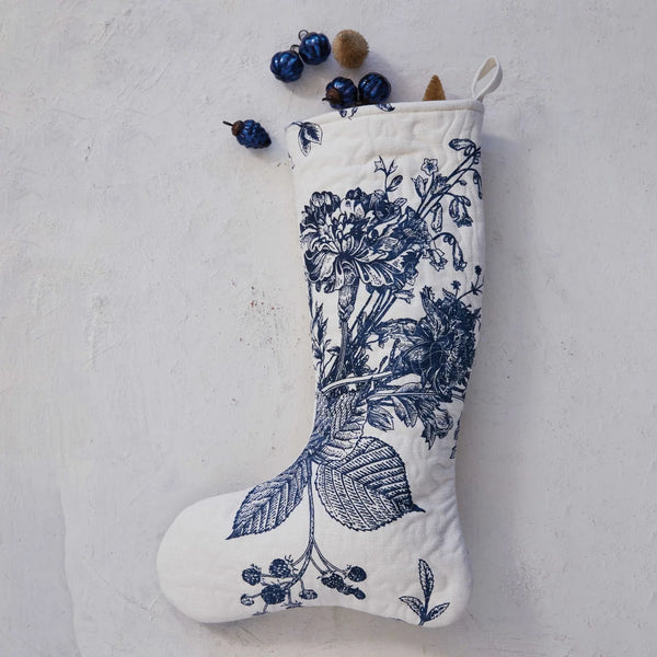A white Christmas stocking with a blue floral botanical print, displayed against a neutral background with a small ornament for scale.
