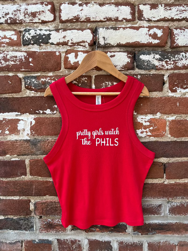 Red tank top with 'pretty girls watch the PHILS' text on a wooden hanger against a brick wall.
