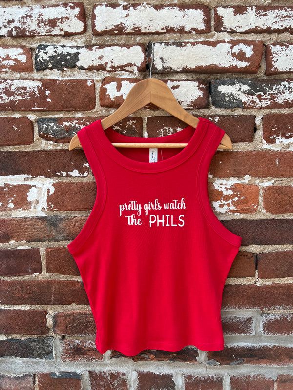 Red tank top with 'pretty girls watch the PHILS' text on a wooden hanger against a brick wall.