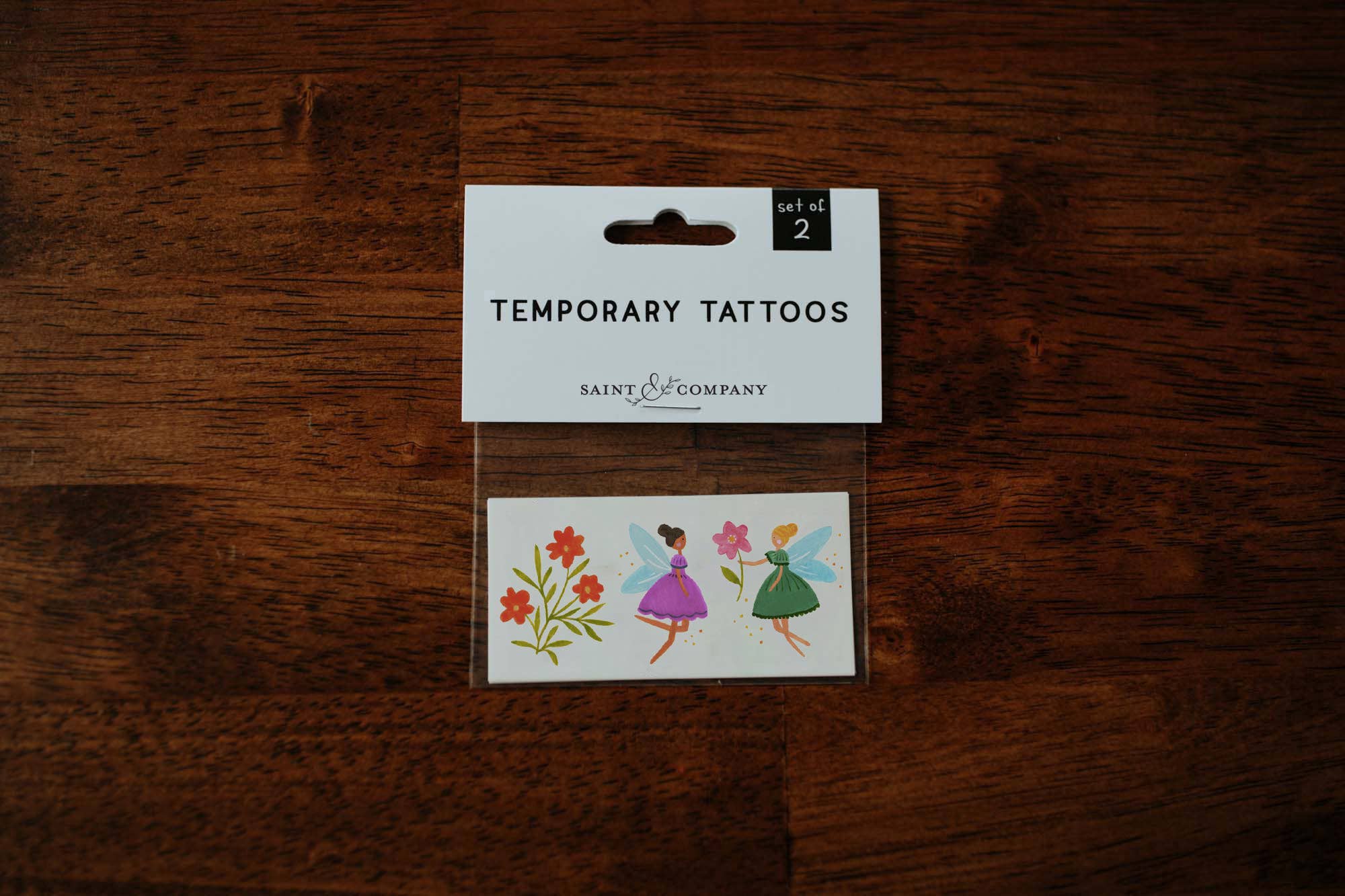 A pack of temporary tattoos featuring two fairies with fairy dust and orange flowers, presented on a wooden surface.