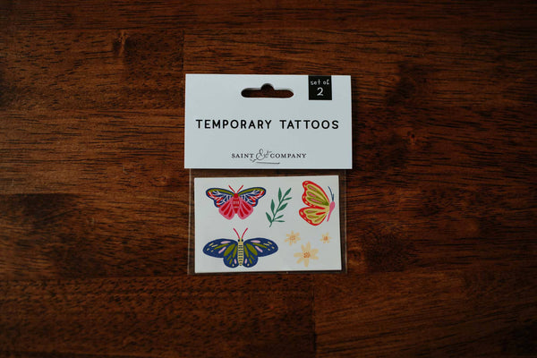 A pack of 'Secret Garden' temporary tattoos featuring hand-painted butterflies, flowers, and greenery, placed on a wooden surface.