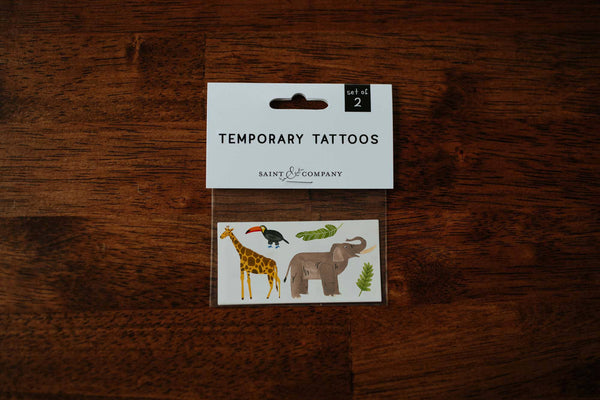 A pack of jungle-themed temporary tattoos featuring illustrations of an elephant, giraffe, and toucan, displayed on a wooden surface.