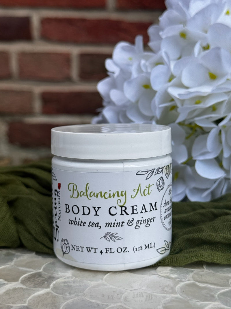 Jar of Balancing Act Body Cream with white flowers and green fabric in the background