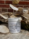 Jar of 'Comfort Body Cream' with vanilla sugar scent on a textured surface with leaves and stones.