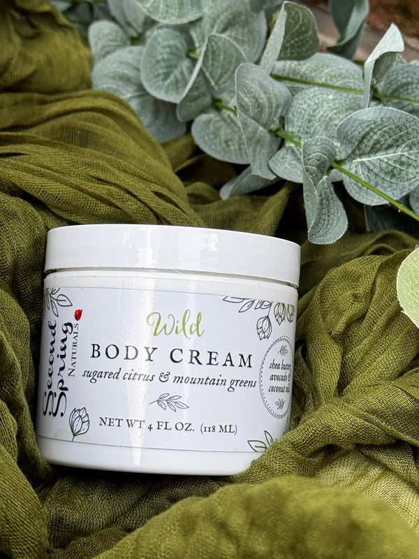 Body cream jar labeled 'Wild' on a green fabric background with eucalyptus leaves.
