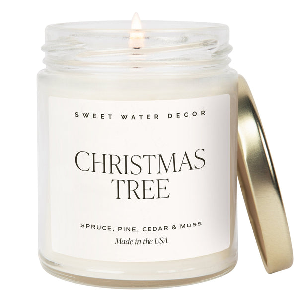 A clear jar candle with a label reading 'Christmas Tree' from Sweet Water Decor, with a lit wick and a metallic lid.