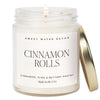 A clear jar soy candle labeled 'Cinnamon Rolls' with a lid and a label that includes the scent notes of cinnamon, icing, and buttery pastry. The candle is placed on a white surface with a lit wick.