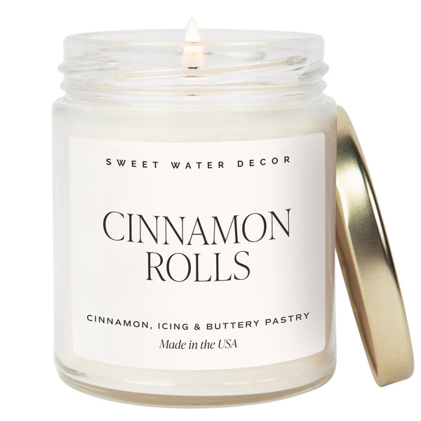 A clear jar soy candle labeled 'Cinnamon Rolls' with a lid and a label that includes the scent notes of cinnamon, icing, and buttery pastry. The candle is placed on a white surface with a lit wick.