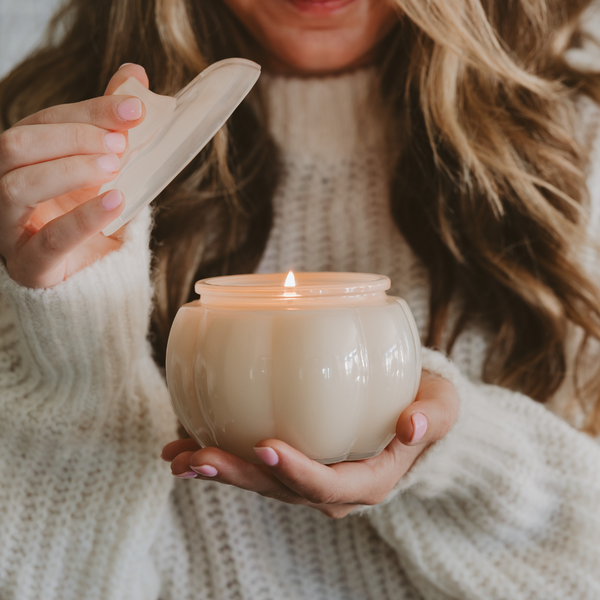 Person holding a lit candle in a white ceramic holder, wearing a beige sweater.
