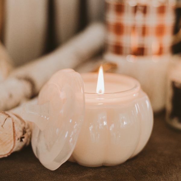 White pumpkin-shaped candle with a lit flame on a brown surface.
