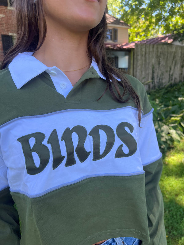 A person wearing a long-sleeve rugby-style shirt with an olive green body, a light blue collar and sleeve sections, and a large dark green 'BIRDS' graphic across the chest, photographed outdoors in a backyard setting.