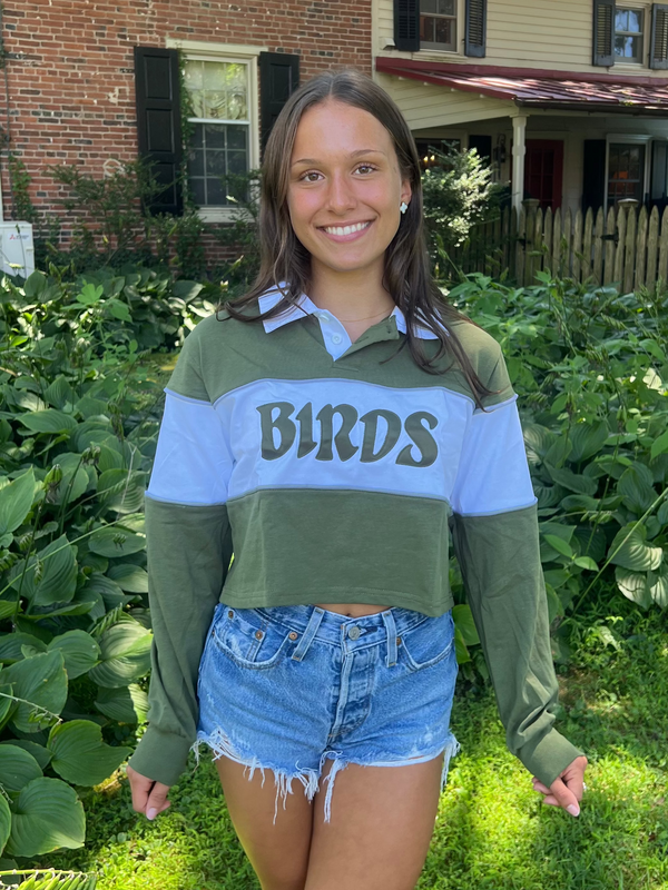 A smiling young woman stands outdoors in a backyard garden wearing a color-blocked long-sleeve crop top with 'BIRDS' printed across the chest in dark lettering, paired with denim shorts.