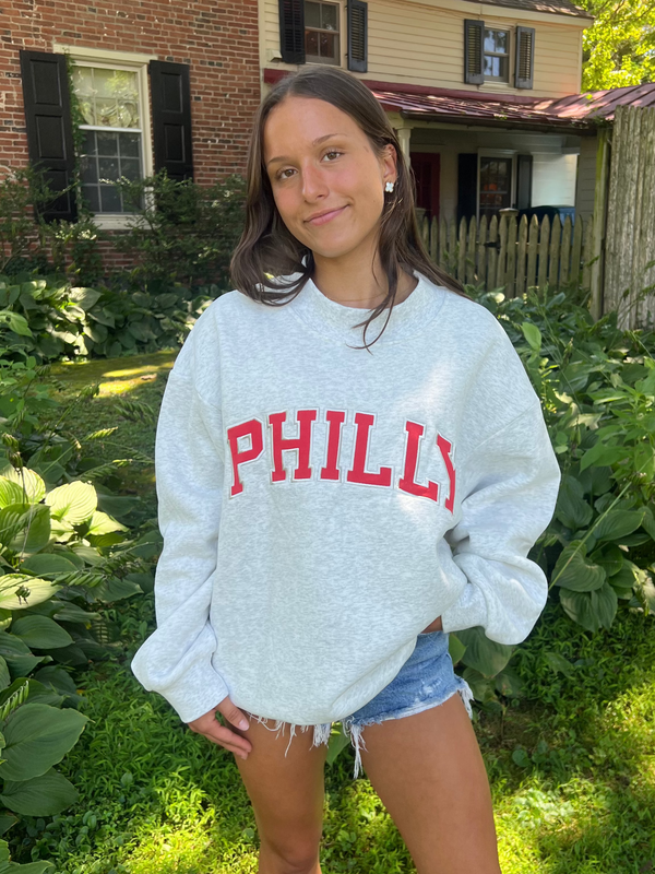 A woman standing outdoors in a grassy yard wearing a light heather gray sweatshirt with 'PHILLY' printed in red across the chest, paired with denim shorts.