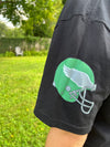 Black shirt with green and white logo on sleeve against a grassy outdoor background