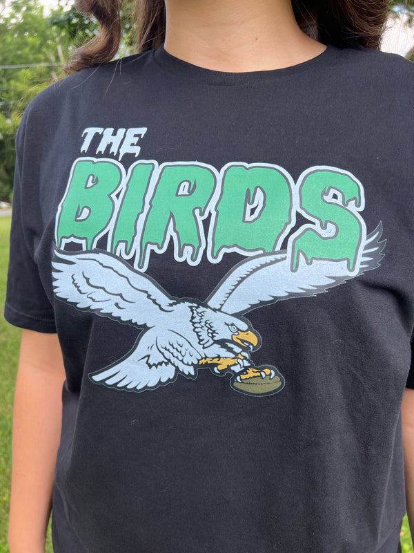 Black t-shirt with green text 'The Birds' and an eagle graphic worn by a person outdoors.