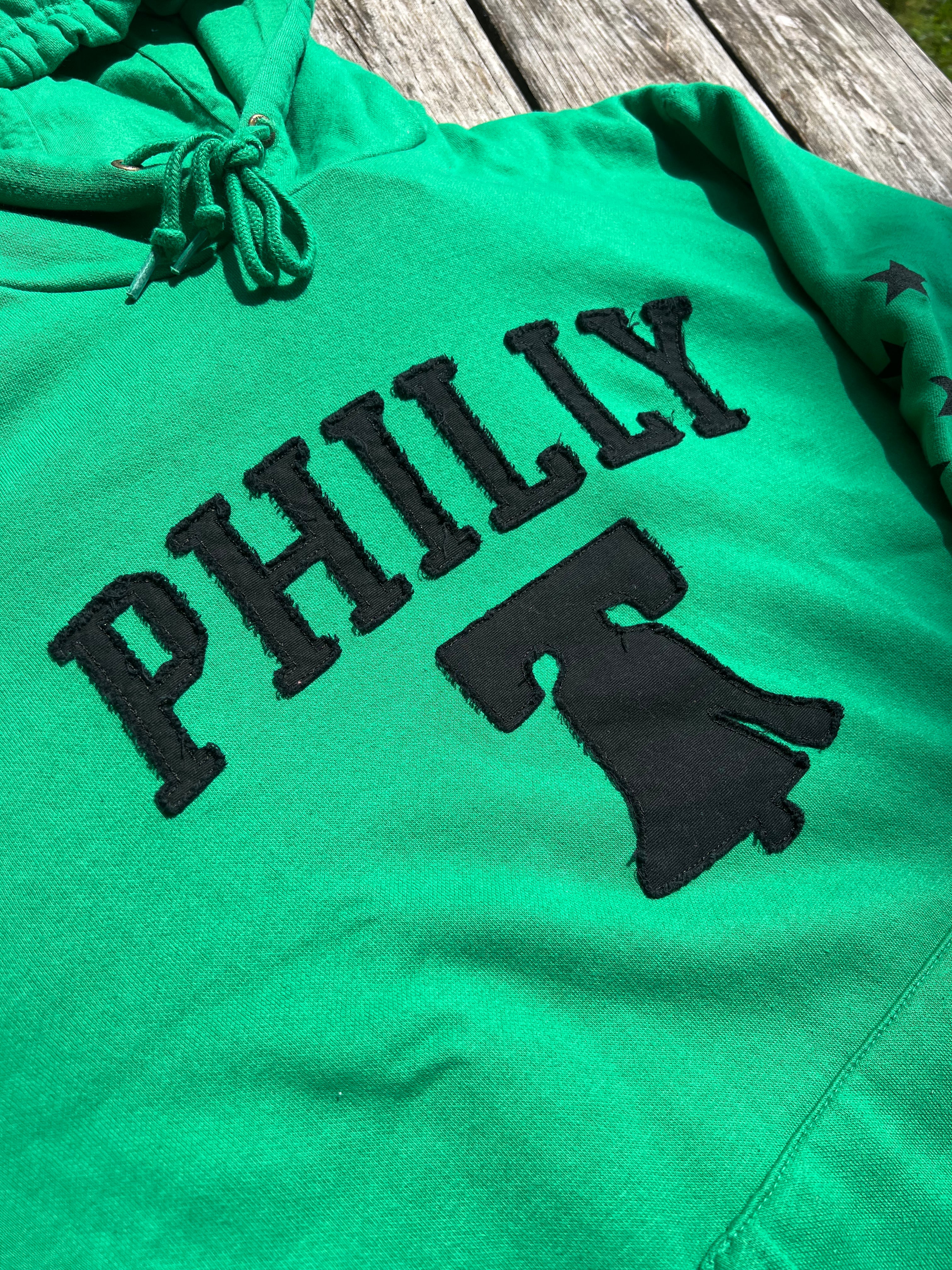 Green hoodie with 'PHILLY' and a bell graphic on a wooden surface