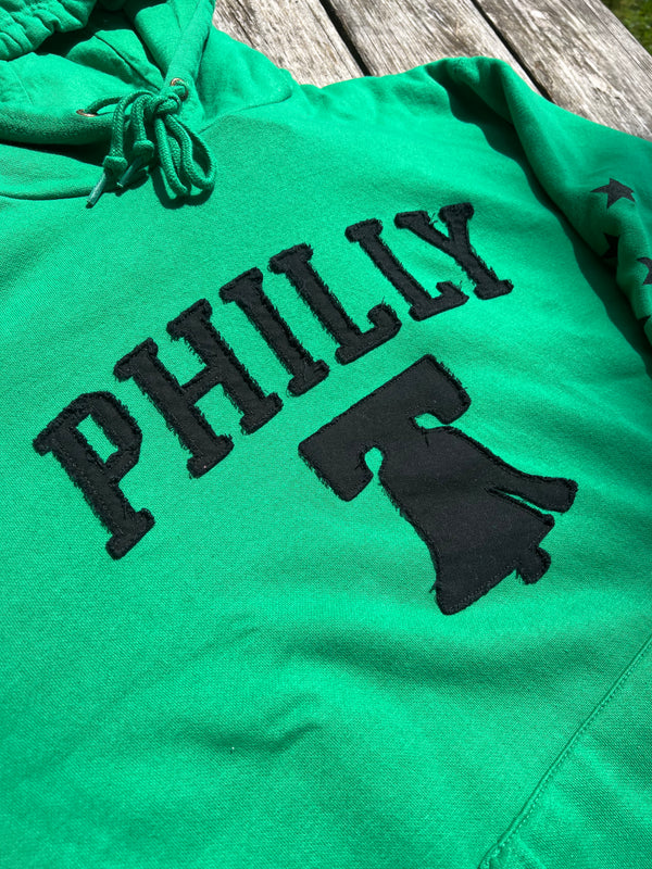 Green hoodie with 'PHILLY' and a bell graphic on a wooden surface