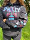 Person wearing a black hoodie with 'PHILLY' and star designs outdoors.