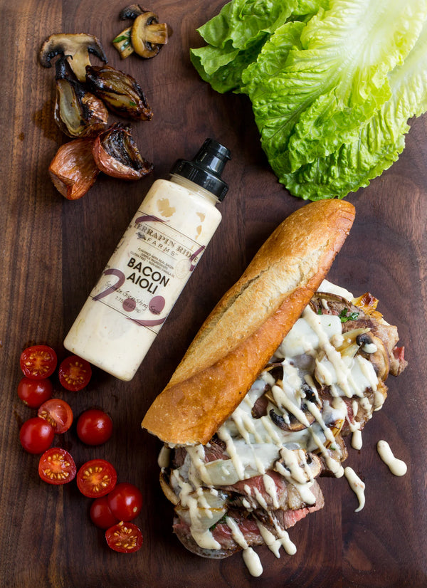 Sandwich with meat and sauce on a wooden board with tomatoes, lettuce, and a bottle of dressing.