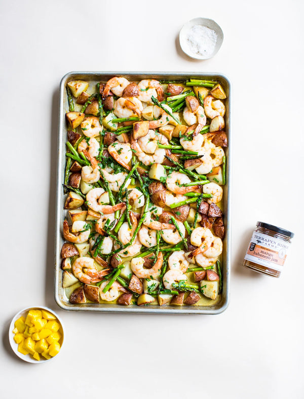 Sheet pan with shrimp, asparagus, and potatoes on a white surface with seasonings.