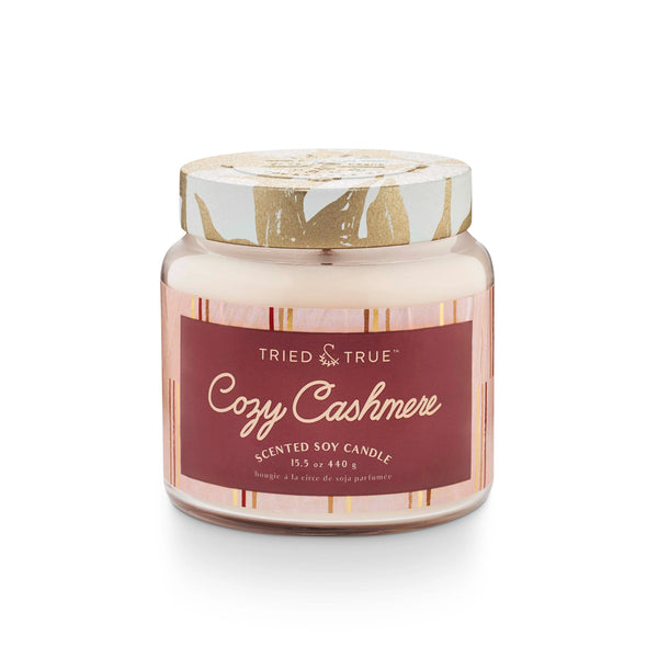 A scented soy candle jar with a label reading 'Cozy Cashmere'. The jar is clear with a white and gold pattern on the lid, and the label has a red background with white text.