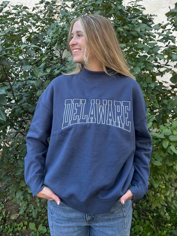 Woman standing outside in a blue mock neck sweatshirt with Delaware stitched on the front in large block letters