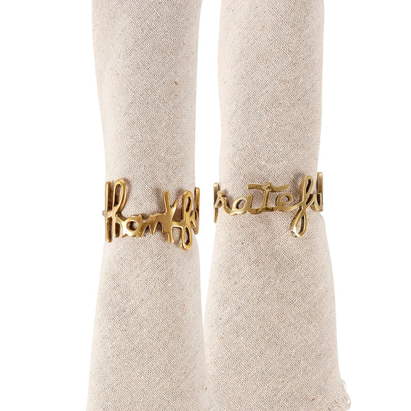 A set of two gold brass napkin rings with the word 'handful of love' written on them, displayed on a textured beige fabric.