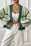 Person wearing a green and white striped cardigan over a white crop top and pants, standing against a white paneled wall.