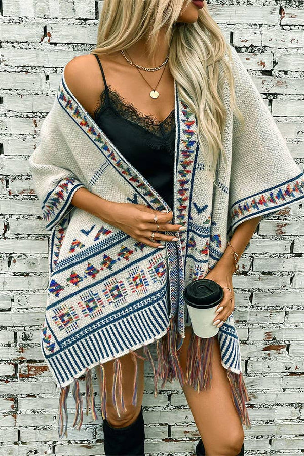 Woman wearing a patterned shawl over a black top, holding a coffee cup against a white brick wall.