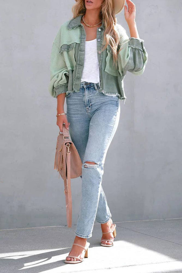 Woman wearing a green jacket, white top, blue jeans, and beige sandals against a gray background.