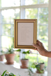 Hand holding a framed poem in front of a window with plants in the background