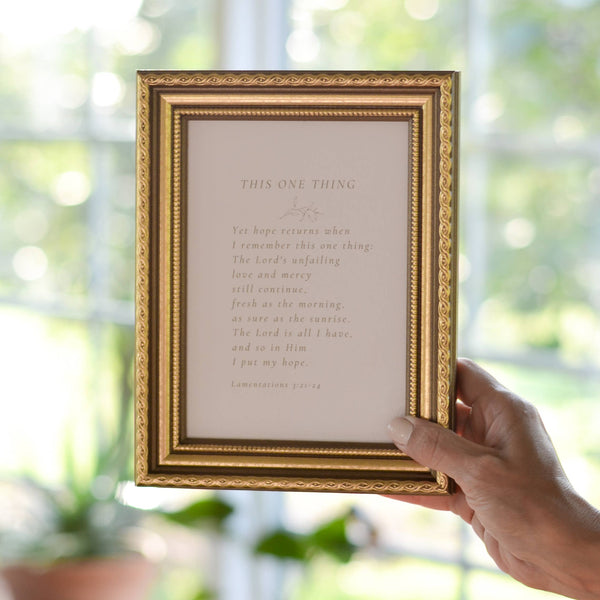 Gold-framed print held by a hand with a blurred greenery background