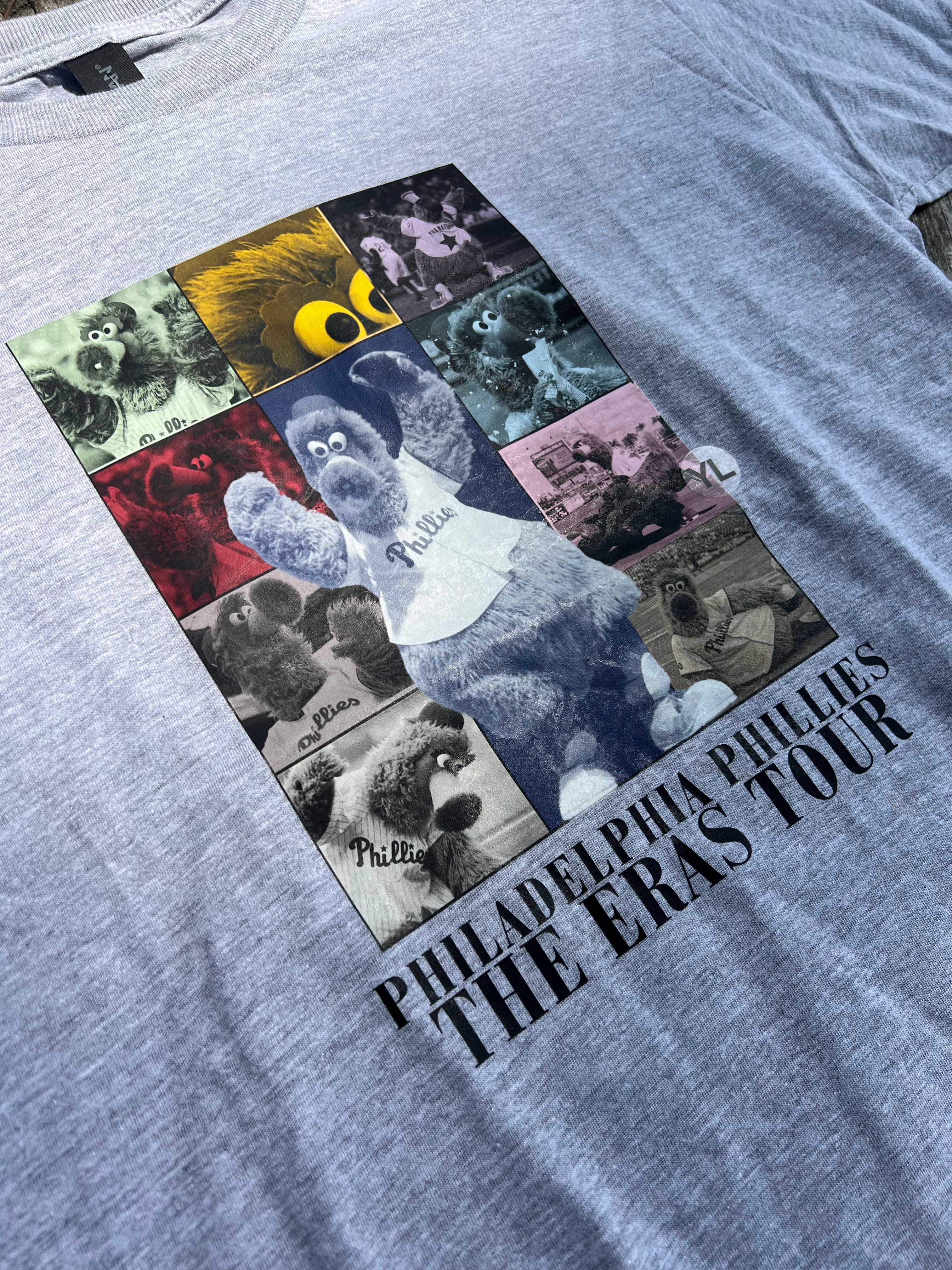 Gray t-shirt with a graphic of a mascot and 'Philadelphia Phillies The Eras Tour' text on a collage background.
