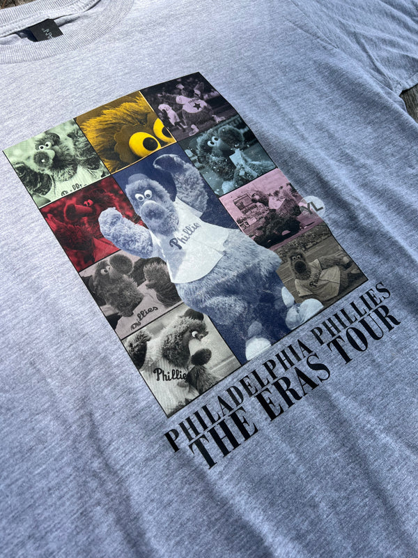 Gray t-shirt with a graphic of a mascot and 'Philadelphia Phillies The Eras Tour' text on a collage background.