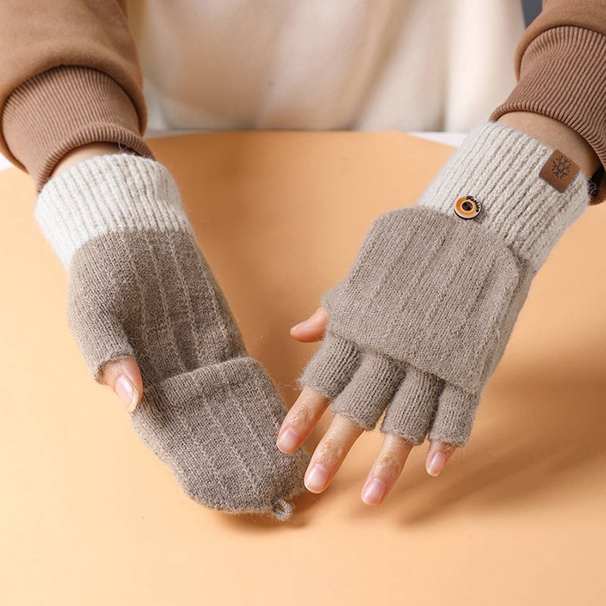 A person wearing a pair of fingerless knit winter gloves with a two-tone design of taupe and cream, featuring a small decorative button on the cuff.