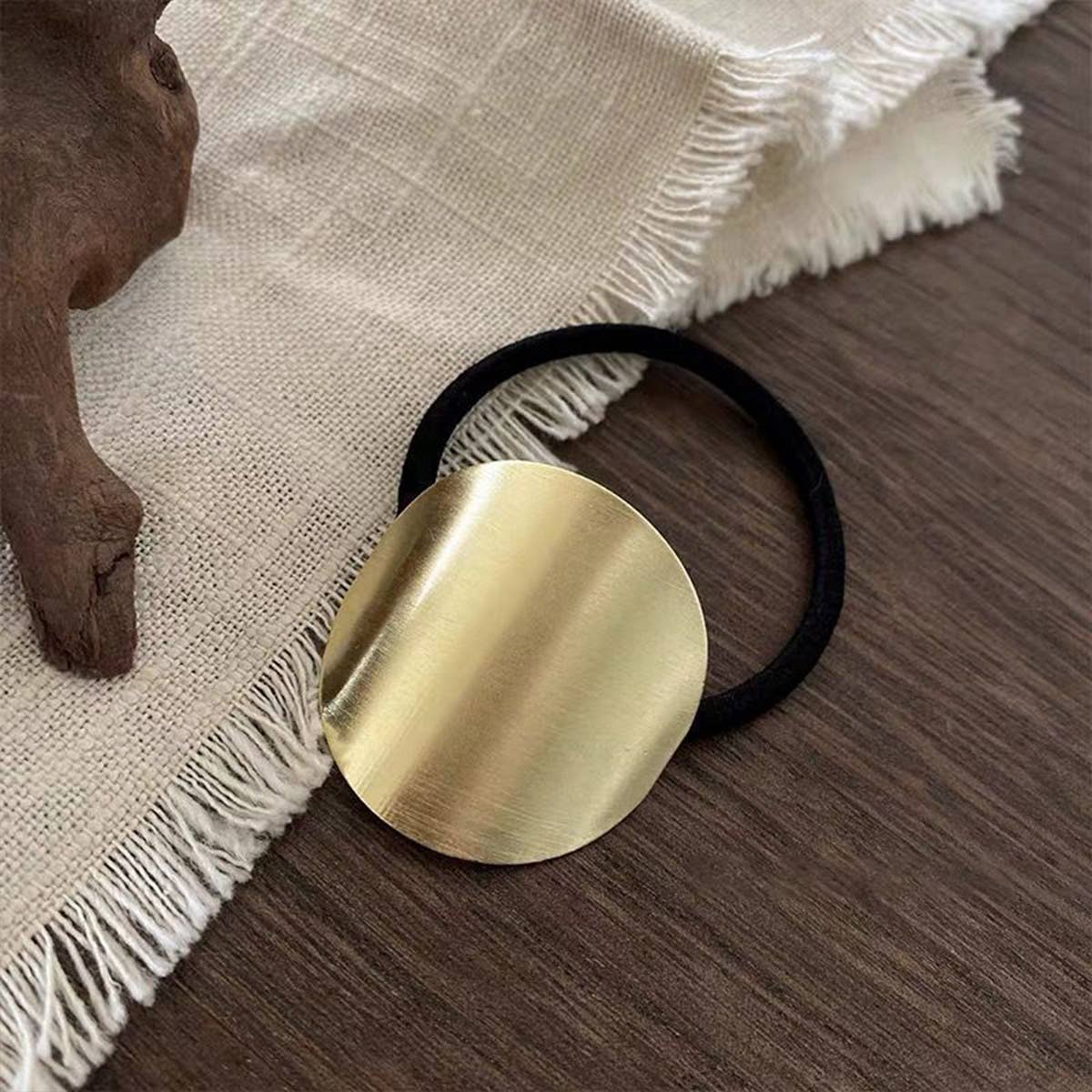 Gold hair tie with a round gold disc on a wooden surface with a textured fabric background