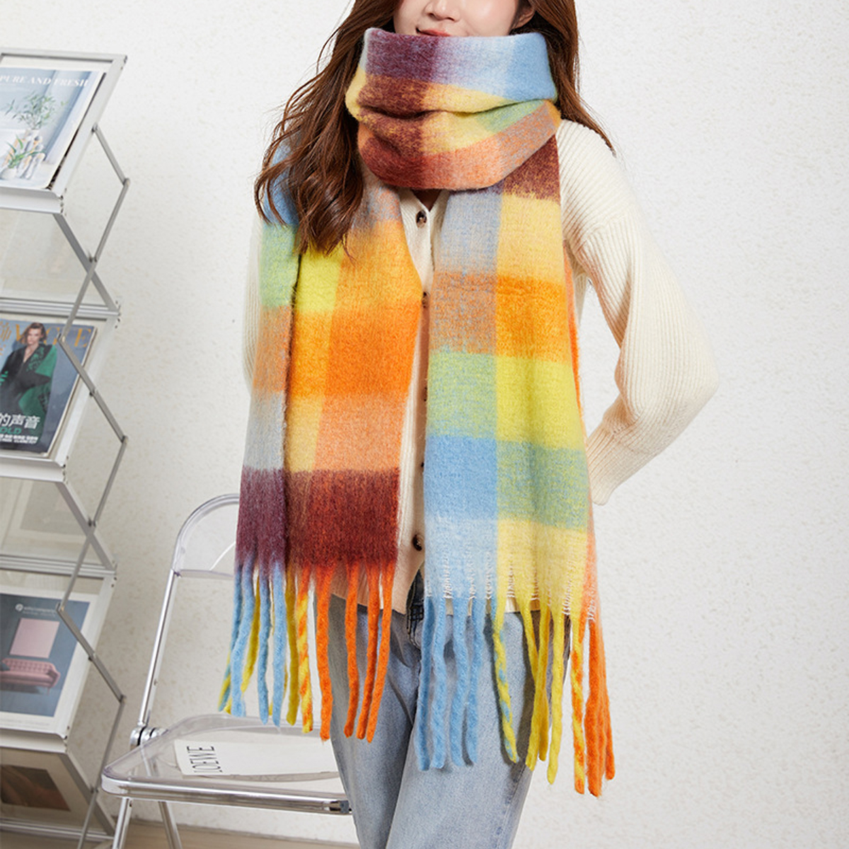 A woman wearing a thick, multicolor plaid scarf with fringe ends, paired with a cream knit sweater and light blue jeans, standing indoors next to a magazine rack and chair.
