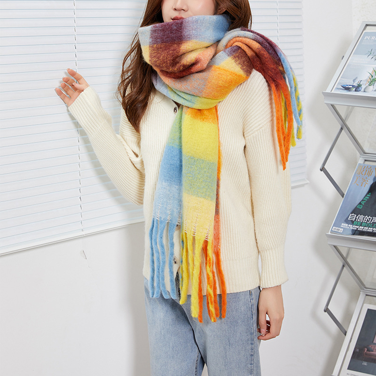 A woman wearing a cream knit sweater and light blue jeans models a thick, multicolored striped scarf with fringe. The scarf features blocks of blue, yellow, orange, brown, and purple.
