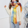 A woman wearing a cream knit sweater and light blue jeans models a thick, multicolored striped scarf with fringe. The scarf features blocks of blue, yellow, orange, brown, and purple.