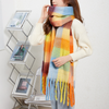 A woman wearing a thick, multicolor plaid scarf with fringe tassels, styled over a cream-colored knit sweater. The scarf features a soft gradient of blue, yellow, orange, green, and brown plaid blocks.