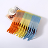A multicolor plaid fringed scarf with orange, yellow, green, and blue sections, styled on a white surface next to a magazine and a small white cup.