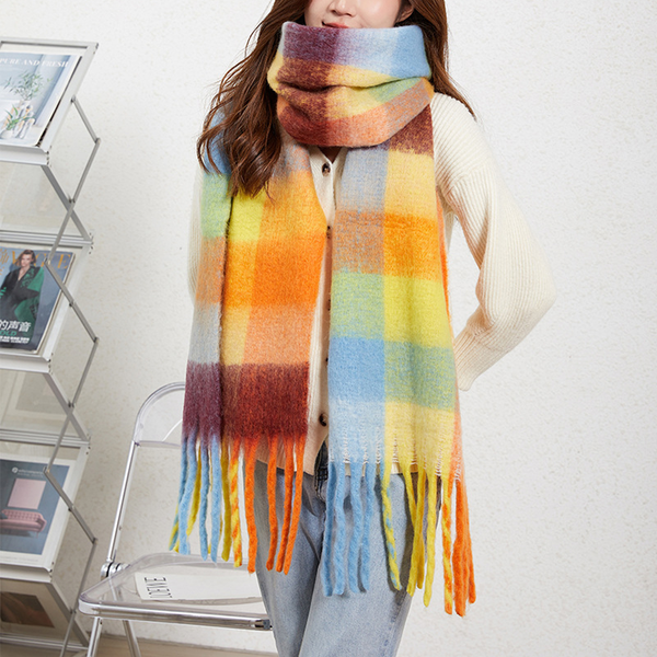 A woman wearing a thick, multicolor plaid scarf with fringe ends, paired with a cream knit sweater and light blue jeans, standing indoors next to a magazine rack and chair.