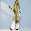 A woman models a thick, textured knit scarf in sage green with orange and white accents, styled with a gray cardigan, white pants, and black sneakers against a light blue studio background.