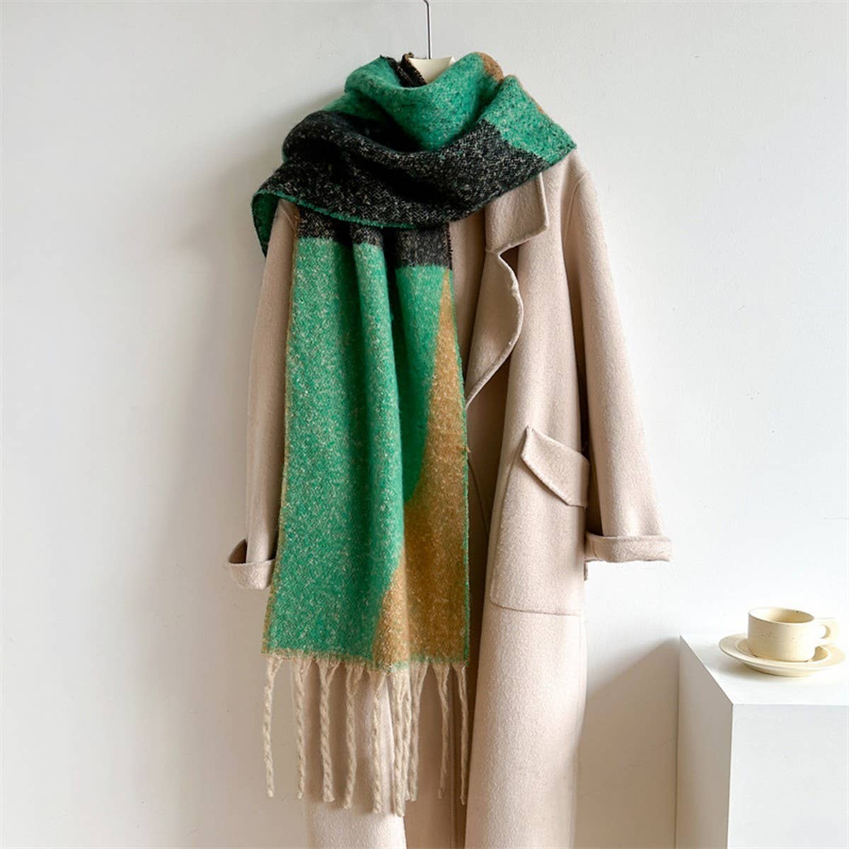 A beige coat displayed on a hanger with a chunky knit scarf draped over it. The scarf features a color-blocked pattern with a bright green section, a black textured section, and a warm tan section, finished with fringe at the bottom.