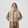 A woman wearing a thick cream-colored knit scarf with multicolored horizontal stripes and fringed ends, styled over a camel coat against a plain light gray background.