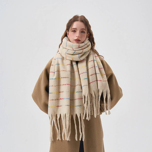 A woman wearing a thick cream-colored knit scarf with multicolored horizontal stripes and fringed ends, styled over a camel coat against a plain light gray background.