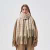 A woman wearing a thick cream-colored knit scarf with thin multicolored horizontal stripes and long fringe ends, styled over a camel-colored coat against a plain light gray background.