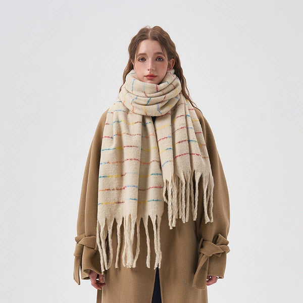 A woman wearing a thick cream-colored knit scarf with thin multicolored horizontal stripes and long fringe ends, styled over a camel-colored coat against a plain light gray background.